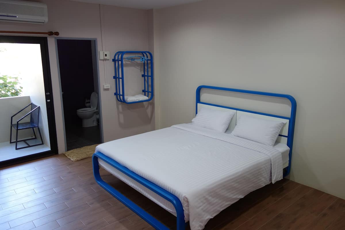 A bedroom with a bed, a blue metal frame, a white mattress, and a blue metal headboard. There is a bathroom door leading to a toilet and a small balcony with a chair.