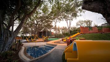 A playground with slides and swings, surrounded by trees and a small pool.
