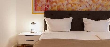 A bedroom with a large bed, two white pillows, a dark headboard, a white bedside table with a lamp, and a colorful abstract painting on the wall.