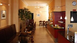 Un vestíbulo de hotel con un mostrador de recepción alargado, un área de descanso con sofá y sillones, una planta grande y un candelabro.