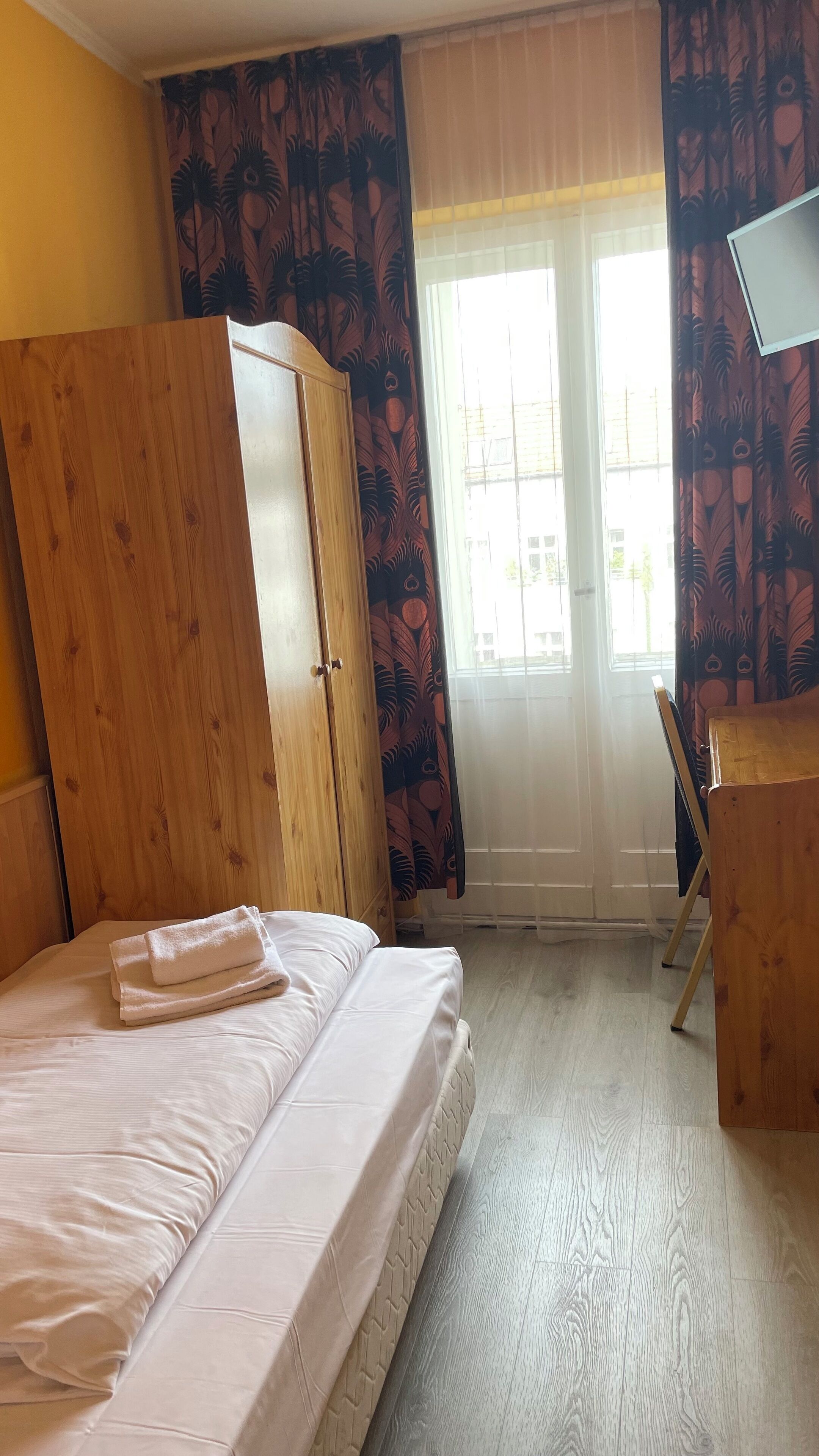 A room with a bed, wooden wardrobe, desk, and a window with curtains.