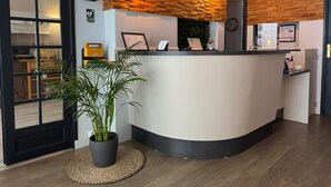 A reception area with a curved counter, wooden wall paneling, hanging light bulbs, a potted plant, and a clock on the wall.