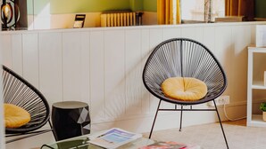 Quarto moderno com mesa de centro de vidro, duas cadeiras de arame pretas, porta-revistas transparente e vista para uma janela com cortinas amarelas.