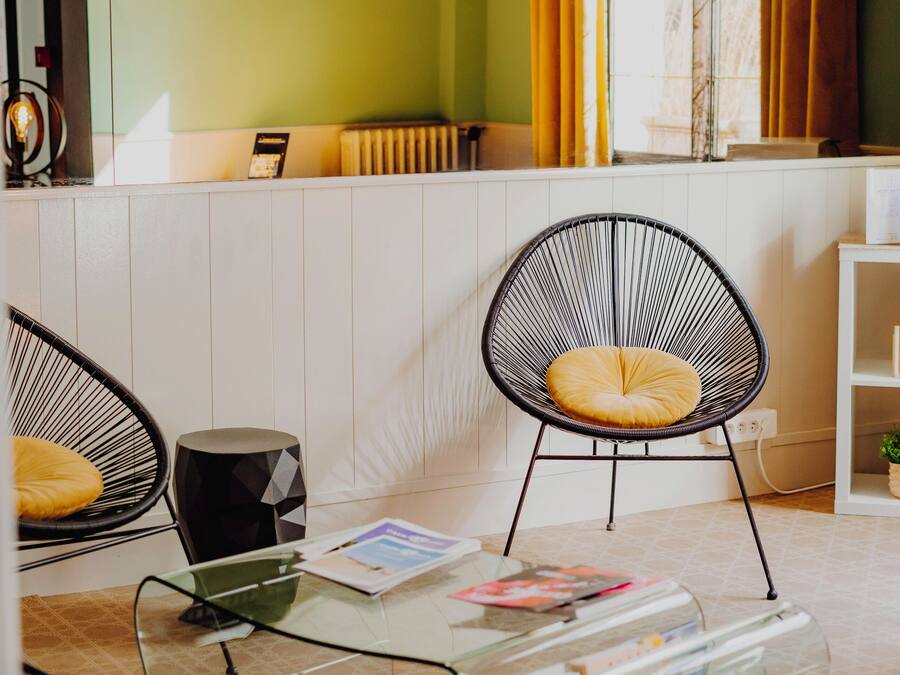 Una habitación moderna con una mesa de centro de vidrio, dos sillas de alambre negras, un atril transparente para revistas y vistas a una ventana con cortinas amarillas.