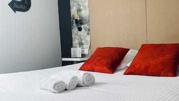 Una cama bien hecha con sábanas blancas y toallas enrolladas, almohadas rojas y un cartel en la pared que dice 'Welcome Home'.