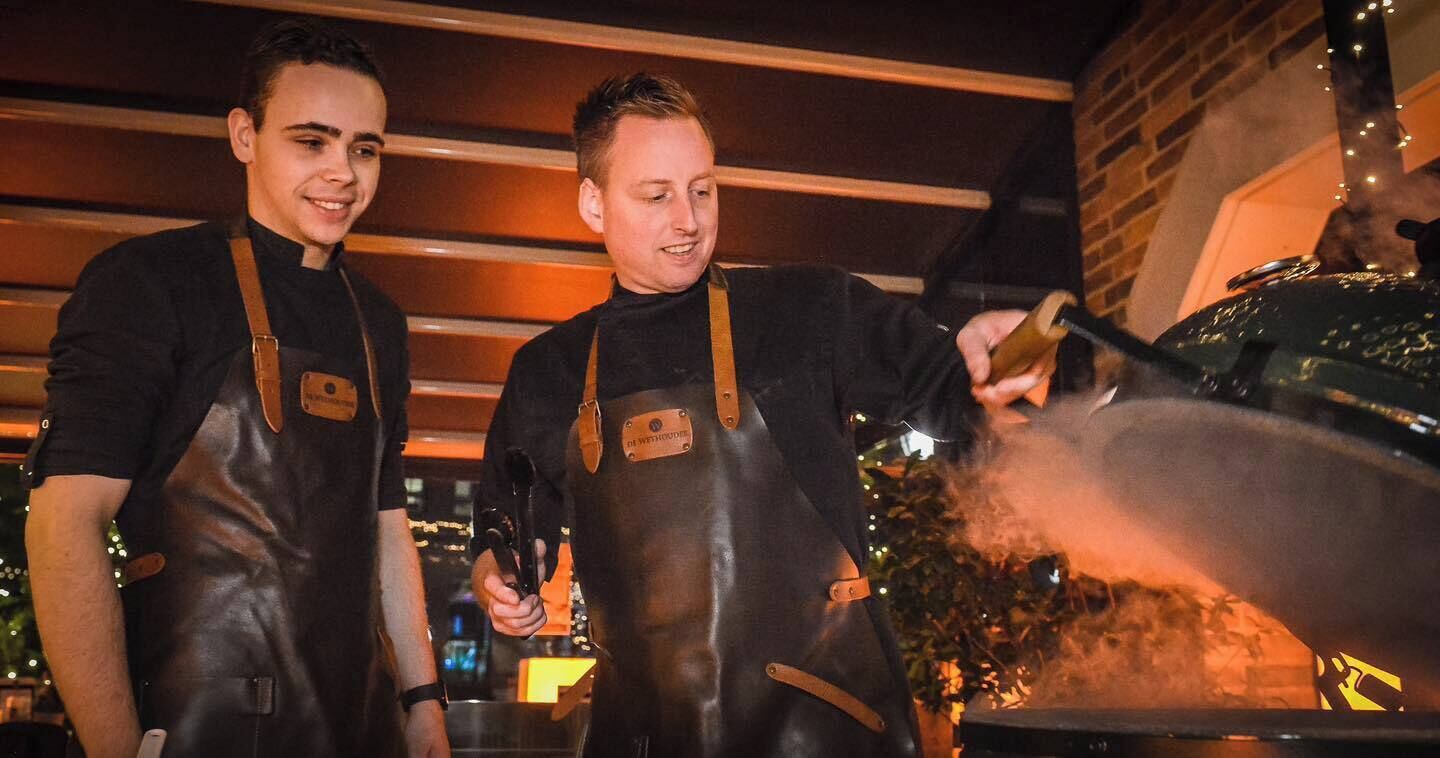 Two individuals in aprons working with a large cooking apparatus outdoors at night.