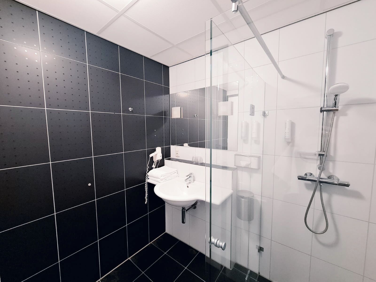 Un baño moderno con cabina de ducha de vidrio, un lavamanos blanco y una pared revestida de baldosas oscuras.