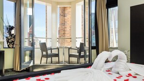 Una camera d'albergo con un letto, un balcone con sedie e vista sugli edifici, e un muro in mattoni.