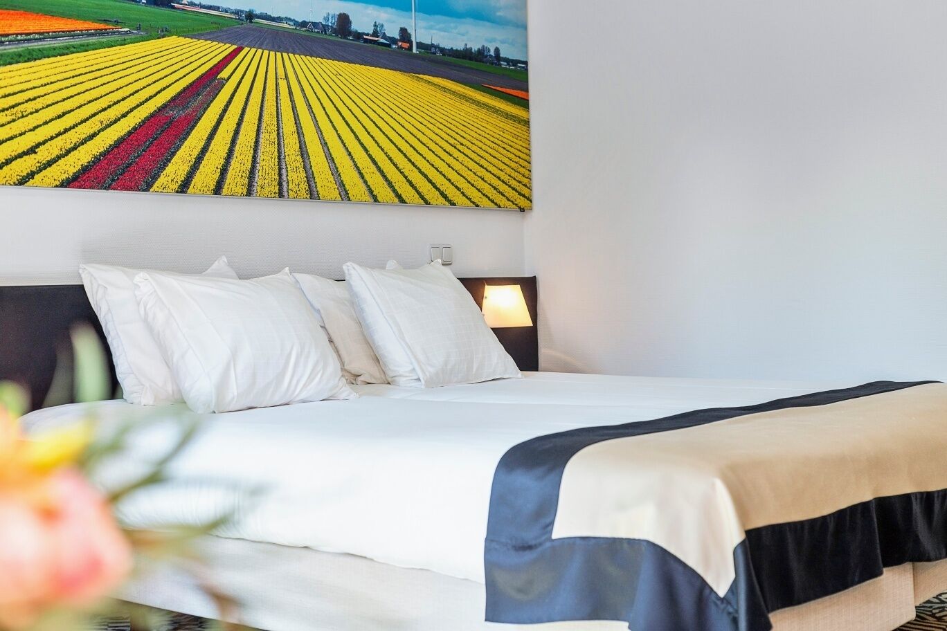 A hotel room with a bed, white pillows, a nightstand with a lamp, and a large painting of a field of flowers on the wall.