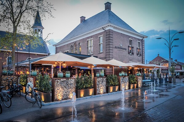 A historic building with a clock tower, outdoor seating with umbrellas, and a water feature. - Hotel de Schout (Denekamp)