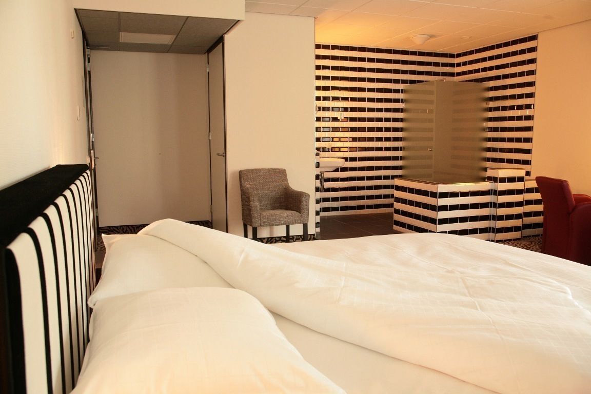 A hotel room with a bed, a chair, a red armchair, and a tiled wall with a unique pattern.