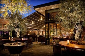 An outdoor dining area with round tables and chairs, surrounded by large potted trees. - Hotel de Schout (Denekamp)