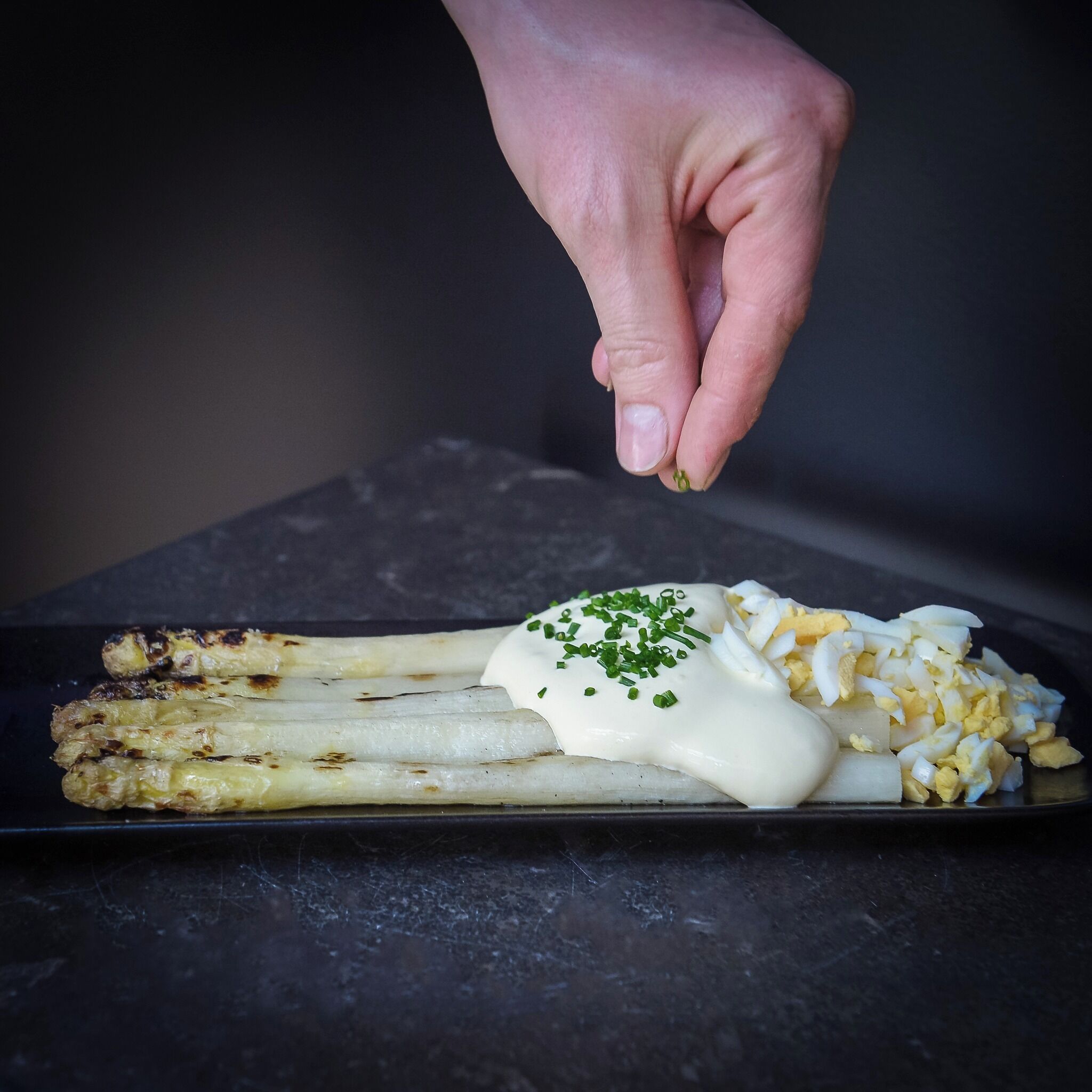 Een hand strooit gehakte groene uien over een gerecht met gegrilde asperges en gekookte eieren.