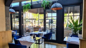 Un'area pranzo interna con tavoli e sedie, ampie finestre e piante pensili.