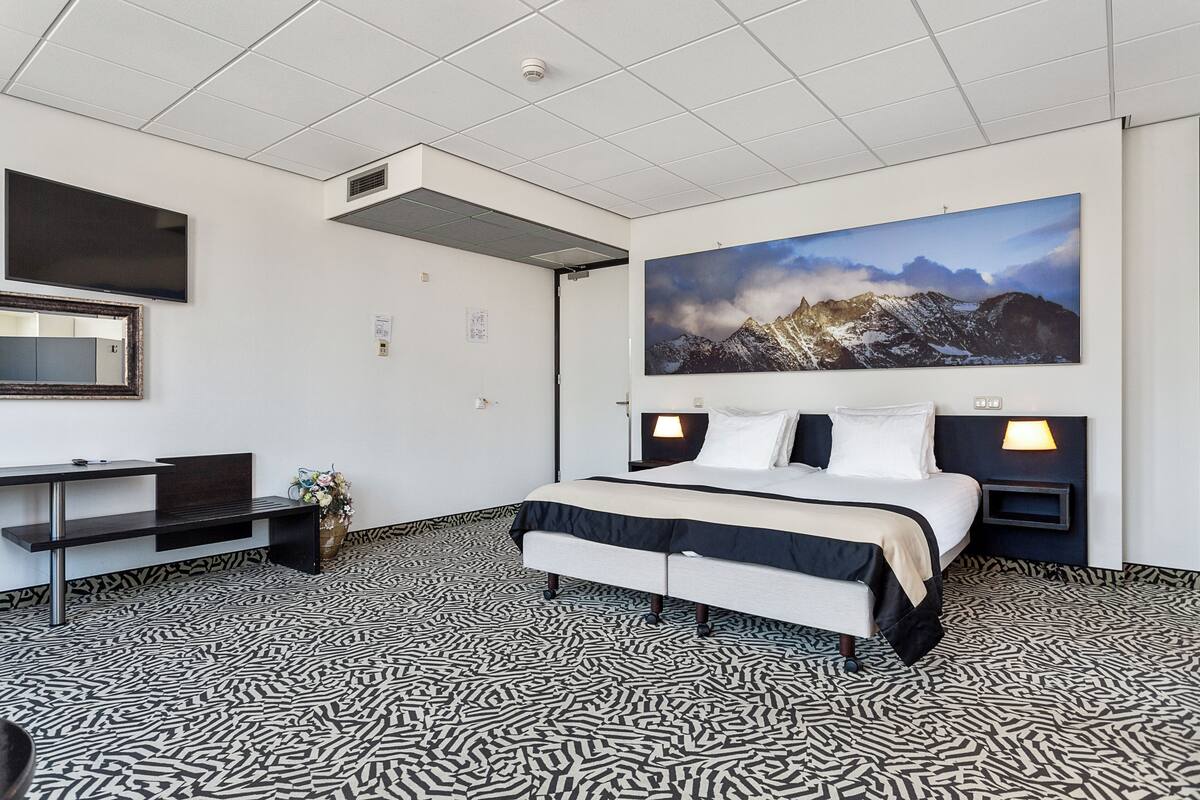 A hotel room with a bed, a desk, a chair, a large painting on the wall, and patterned flooring.