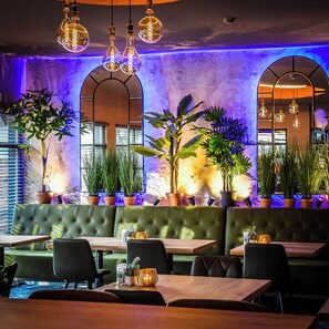 A restaurant interior with a green upholstered booth seating, tables, hanging light fixtures, and potted plants.
