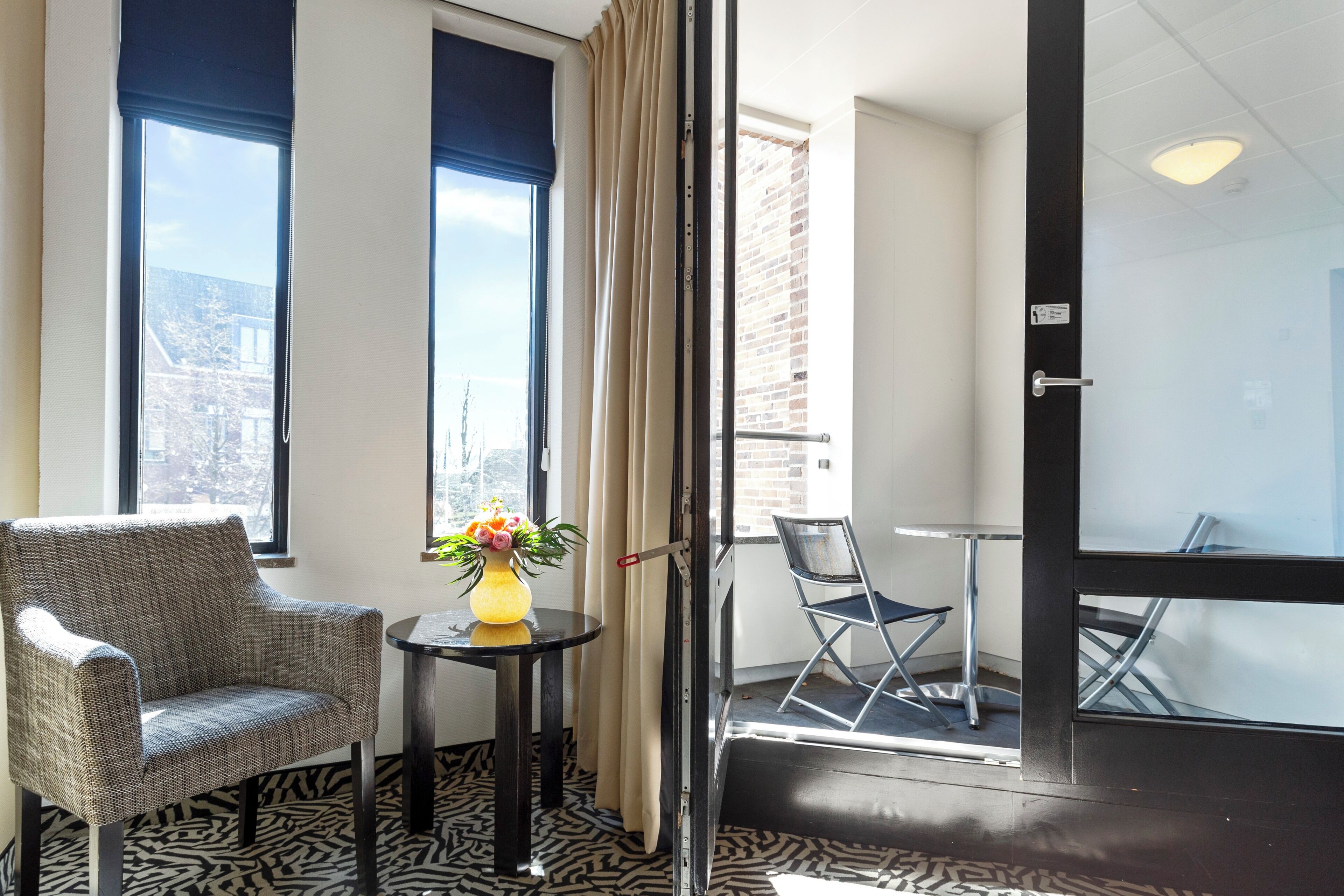 Una habitación con una silla, una mesa pequeña con un florero y vistas a la ciudad a través de amplios ventanales.