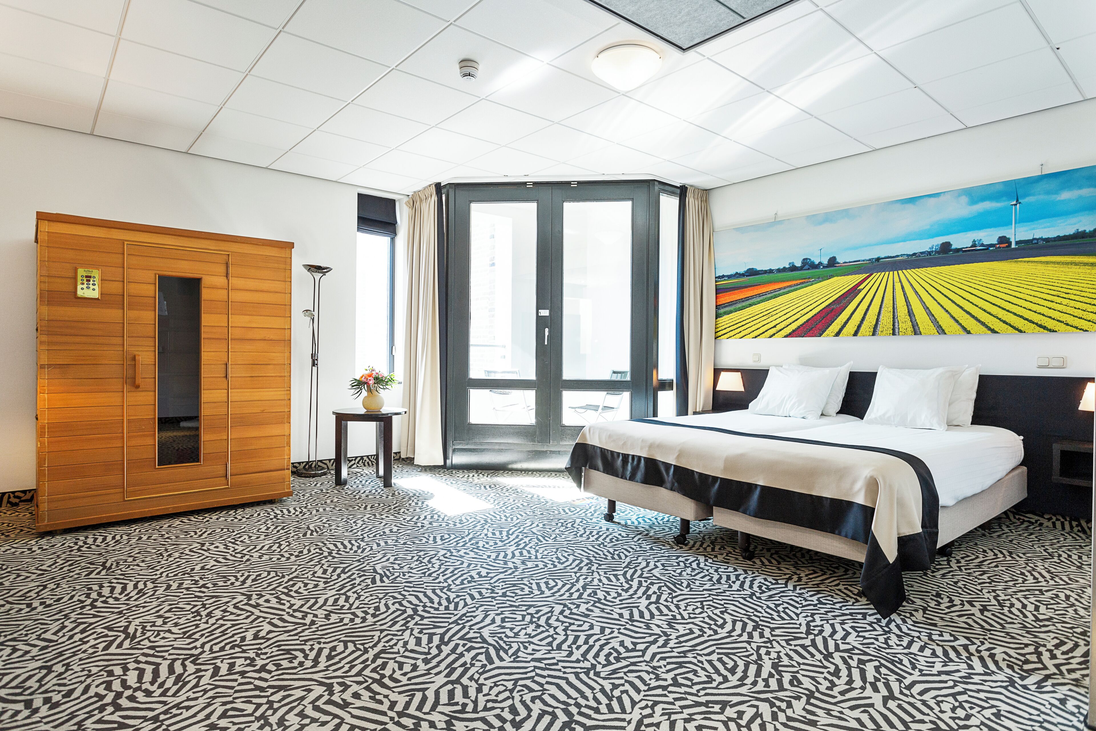 A hotel room with a bed, a wooden wardrobe, a small table with a vase, a painting of a field, and a patterned carpet.