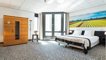 A hotel room with a bed, a wooden wardrobe, a small table with a vase, a painting of a field, and a patterned carpet.