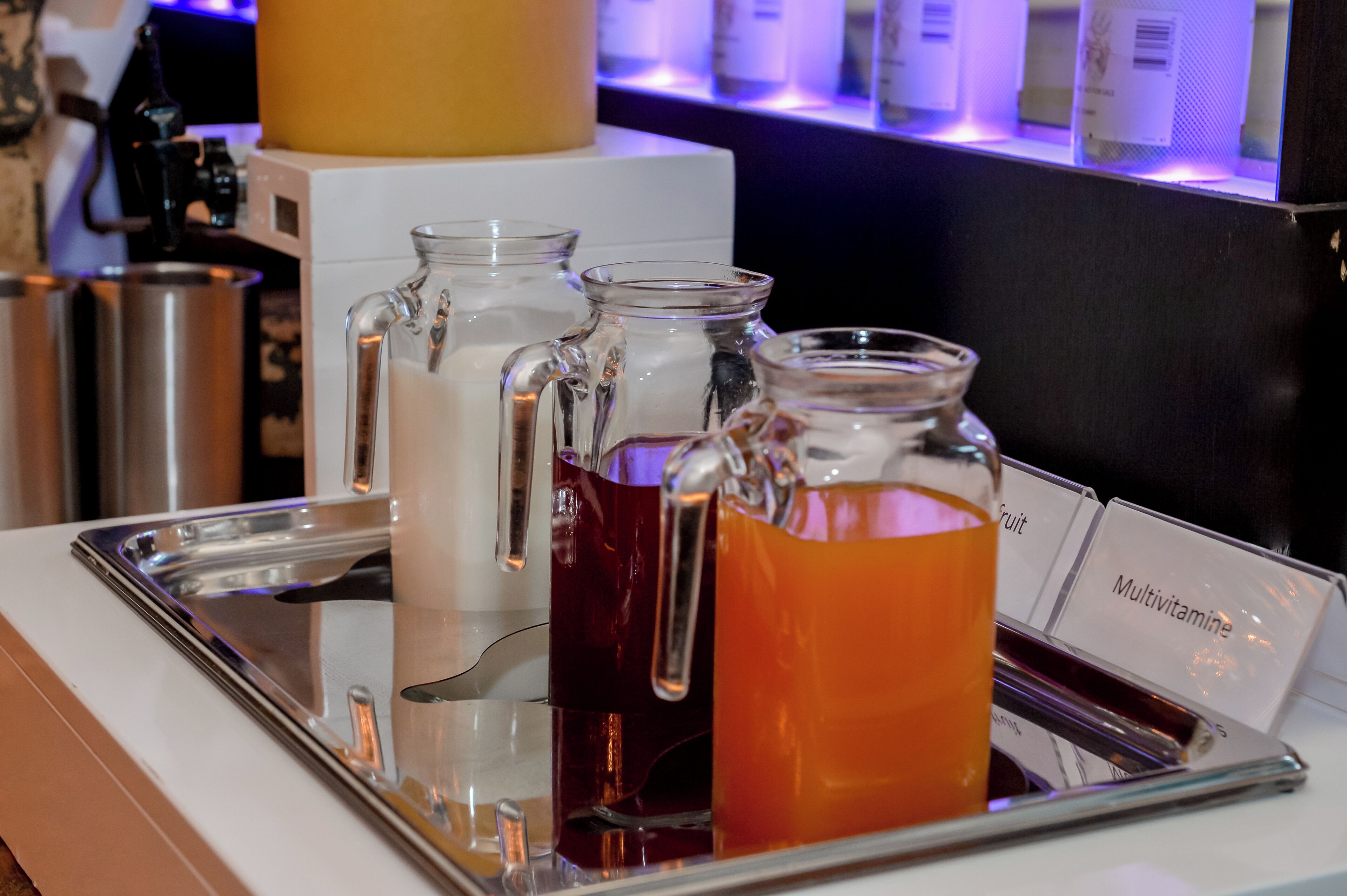 Una estación de bebidas con tres recipientes de vidrio etiquetados 'Fruit', 'Multivitamina' y 'Té'.