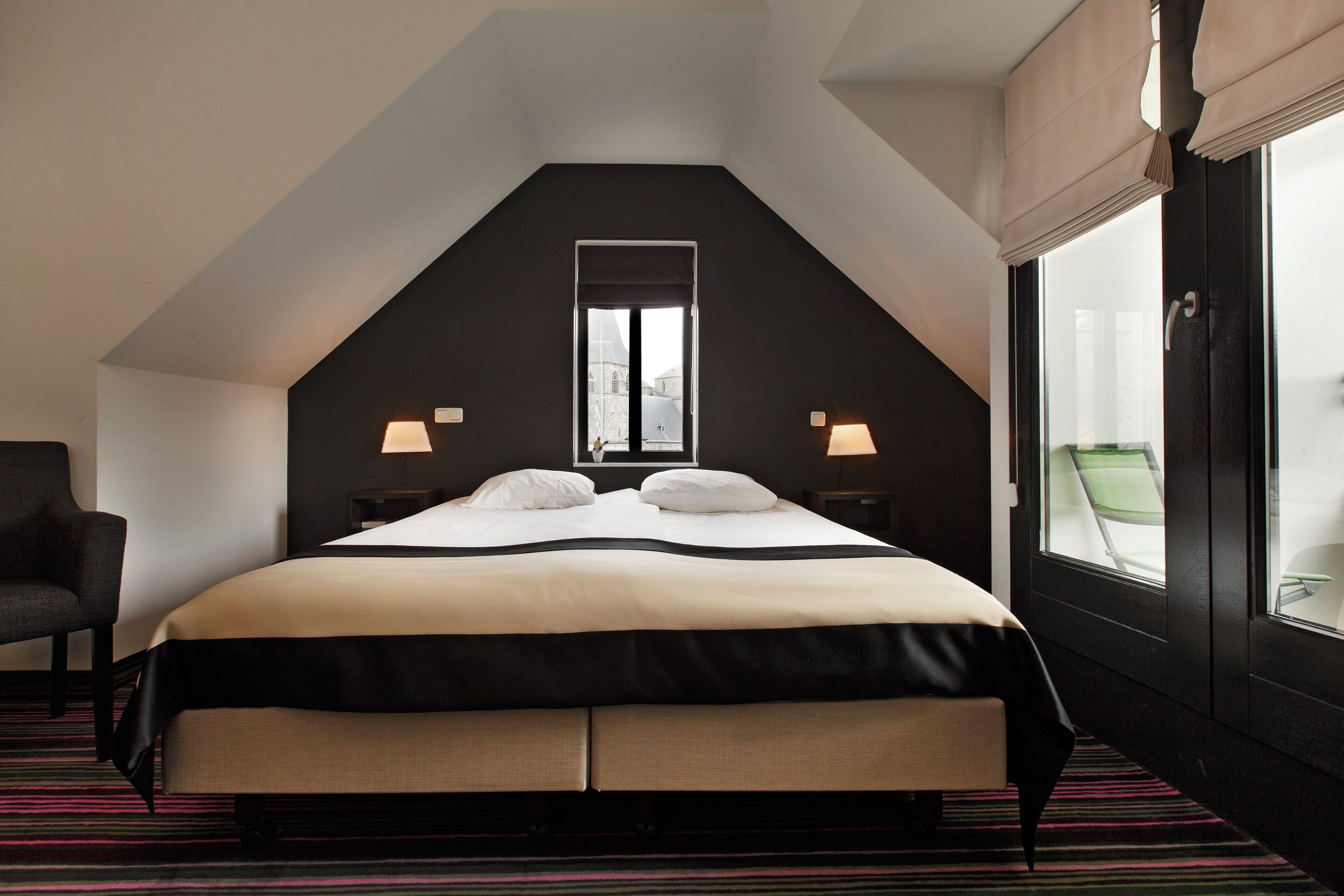 A hotel room with a large bed, two bedside lamps, a window with curtains, a chair, and a view of the cityscape.