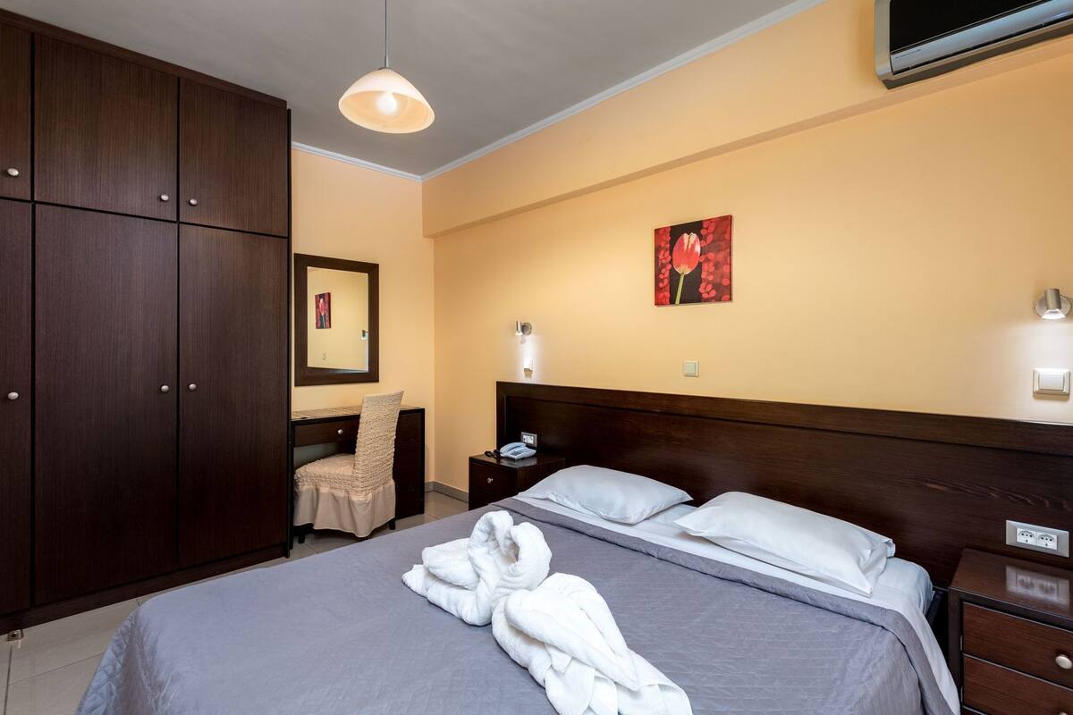 Habitación de hotel con cama, una silla, escritorio, mesita de noche, armario y un cuadro en la pared.