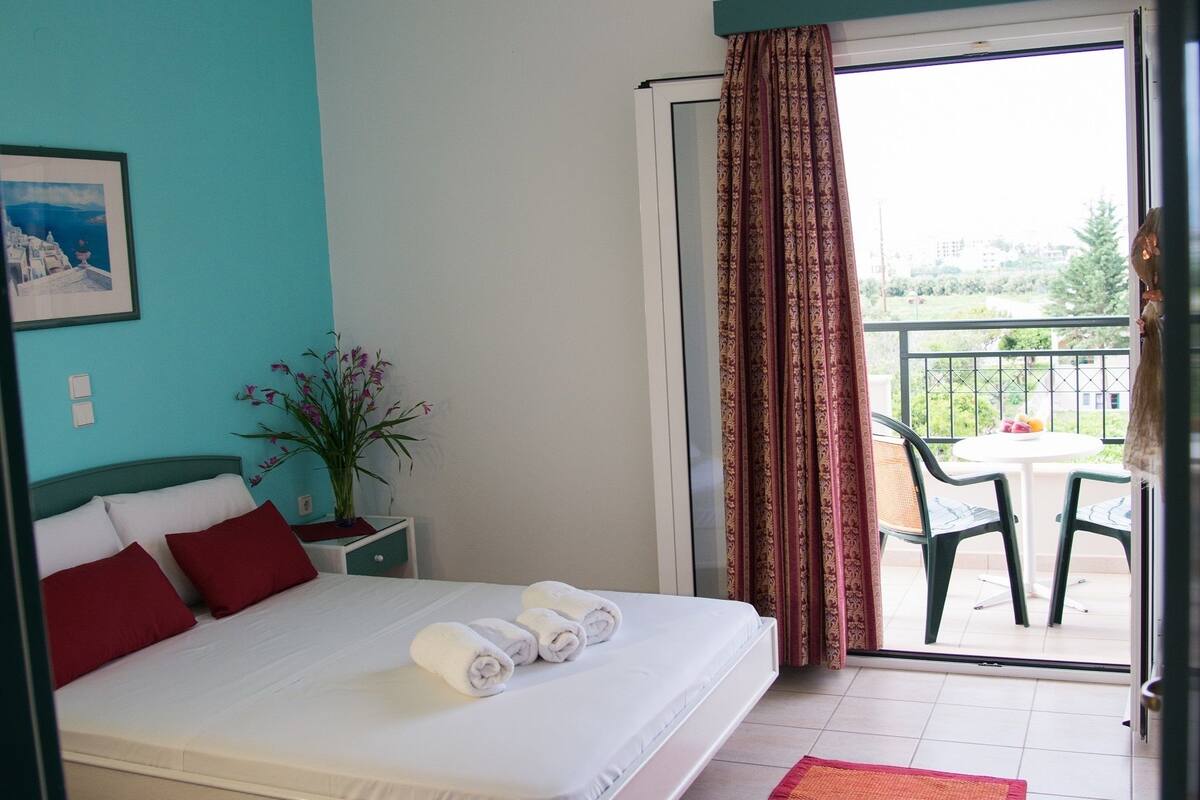 A hotel room with a bed, a potted plant, a balcony with chairs, and a view of the outside.