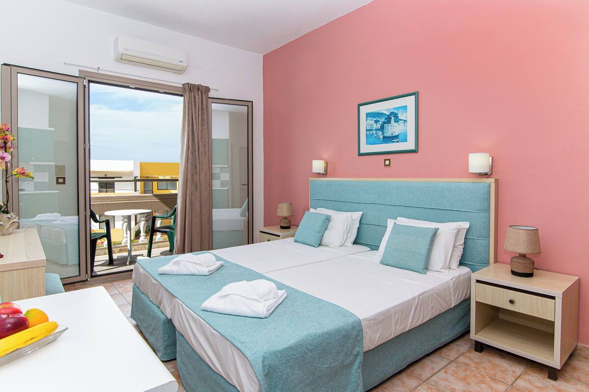 A hotel room with a bed, a desk with a chair, a small table, and a balcony with a table and chairs.