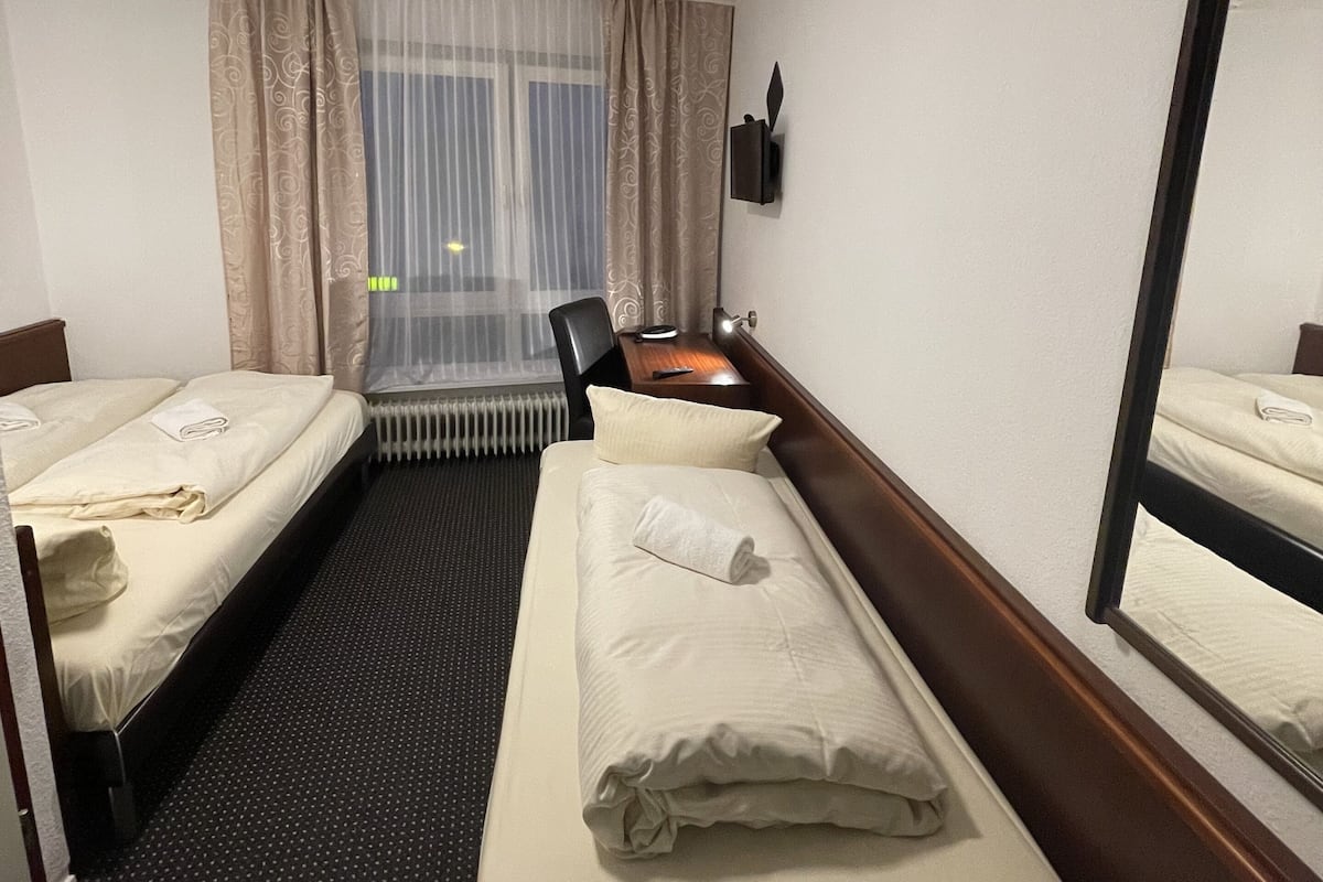 Une chambre d’hôtel avec deux lits simples, un bureau avec une chaise, un radiateur, une fenêtre avec des rideaux légers et un téléviseur fixé au mur.