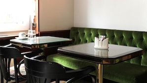 A restaurant interior with a green upholstered booth, two glass-top tables, black chairs, and a wall-mounted light fixture. - Brunnen Hotel (Essen)