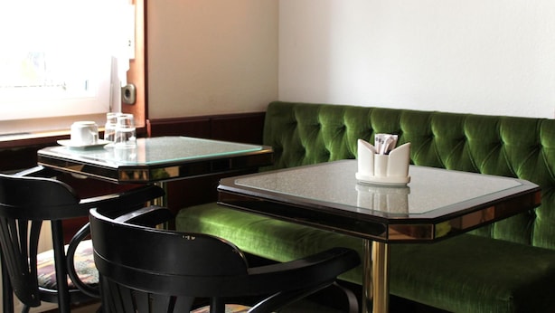A restaurant interior with a green upholstered booth, two glass-top tables, black chairs, and a wall-mounted light fixture. - Brunnen Hotel (Essen)