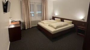 A hotel room with a large bed, a desk, a chair, a radiator, and a window with curtains. - Brunnen Hotel (Essen)