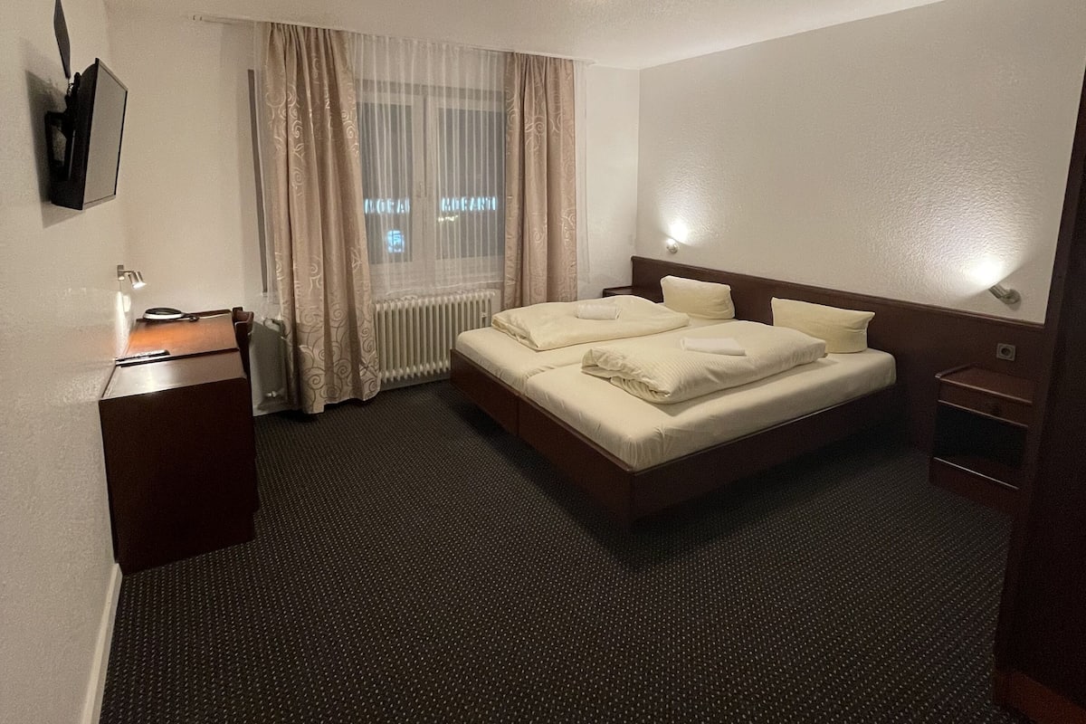 Une chambre d’hôtel avec un grand lit, un bureau, une chaise, un radiateur et une fenêtre avec des rideaux.