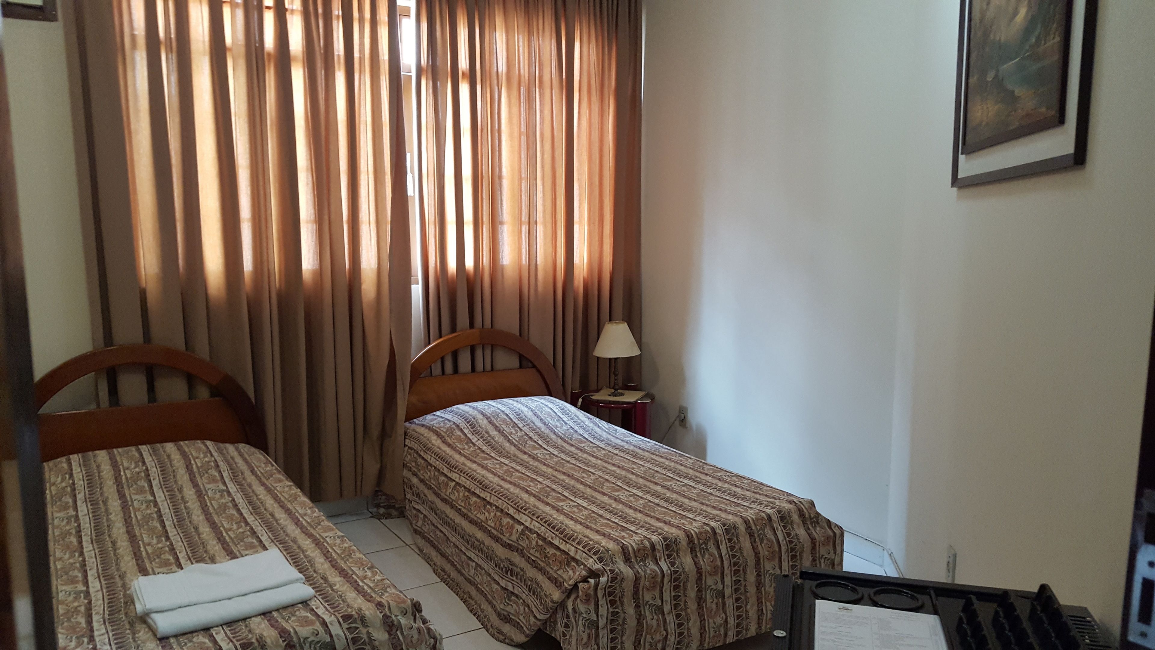 A small hotel room with two single beds, a nightstand with a lamp, and a painting on the wall.