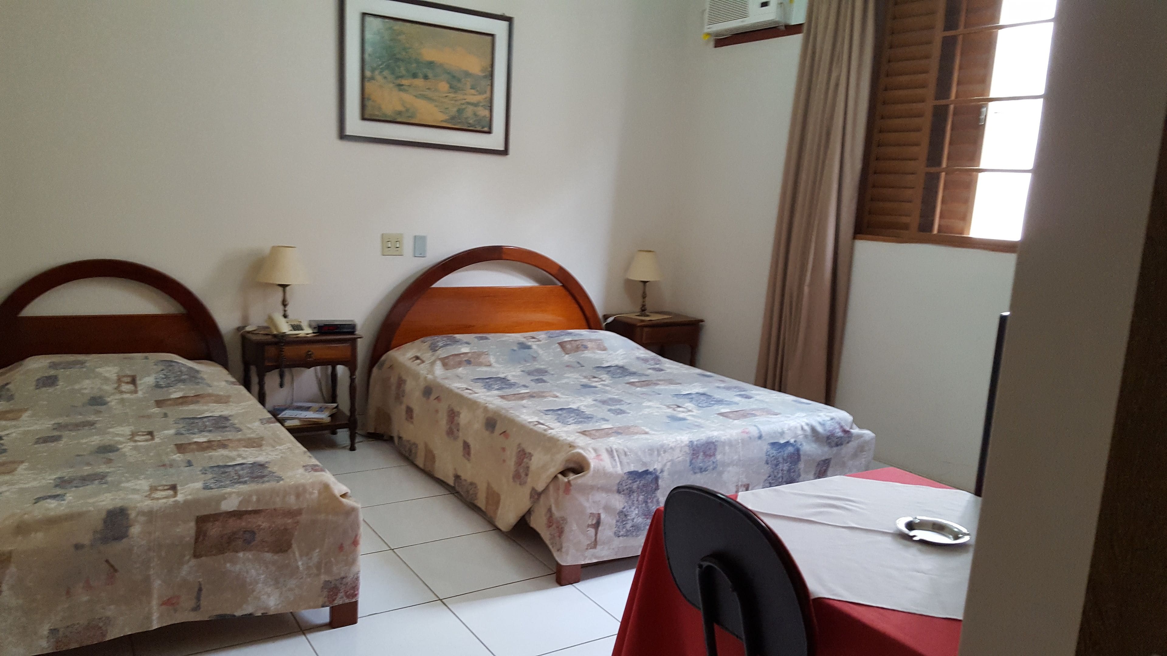 Quarto com duas camas de solteiro, uma pequena mesa com uma cadeira, um quadro na parede e uma janela com cortinas.