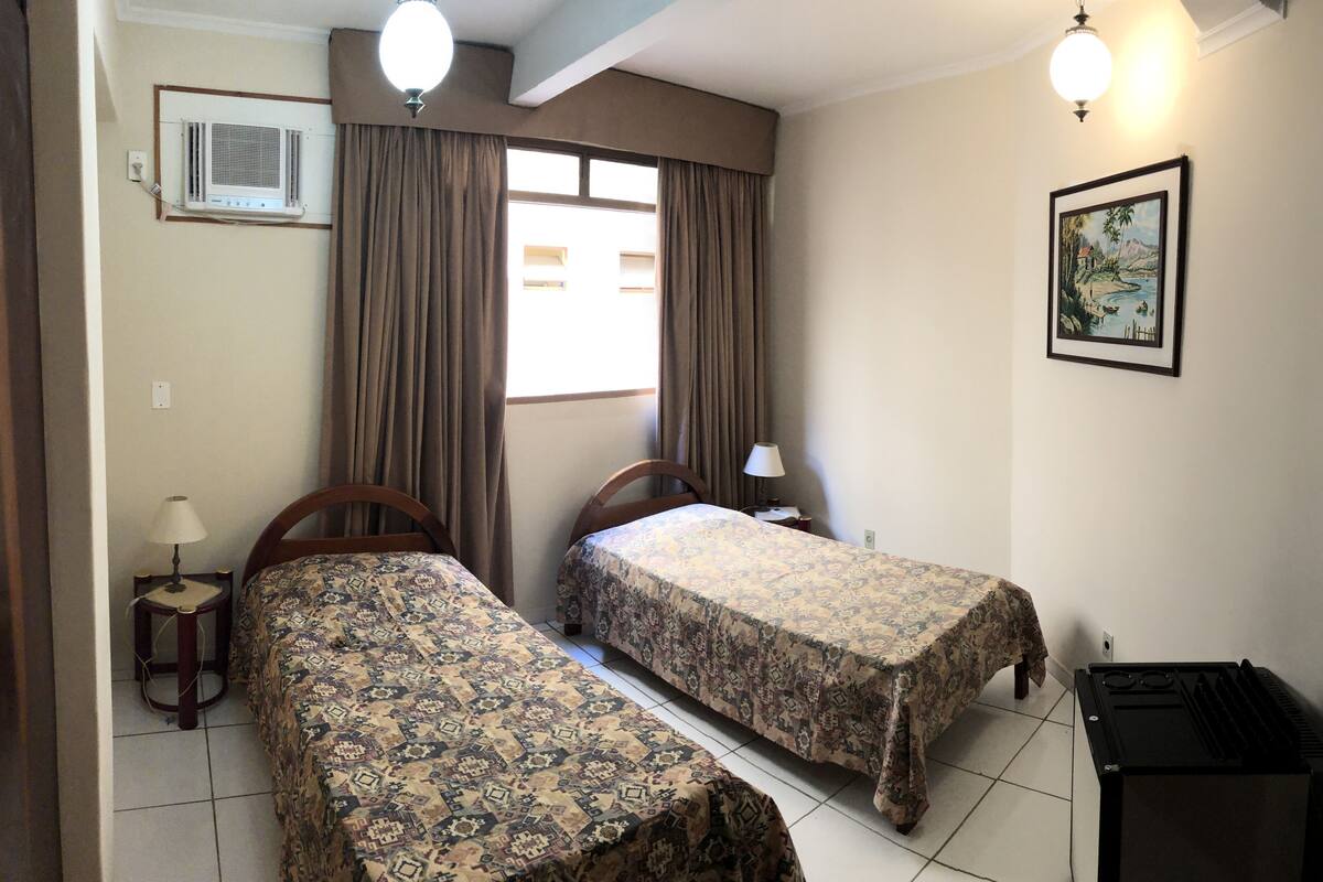 Une chambre avec deux lits, une fenêtre avec des rideaux, un climatiseur fixé au mur, une petite table avec une lampe et un tableau encadré accroché au mur.