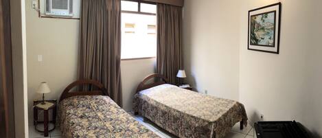 A room with two beds, a window with curtains, a wall-mounted air conditioner, a small table with a lamp, and a framed picture on the wall.