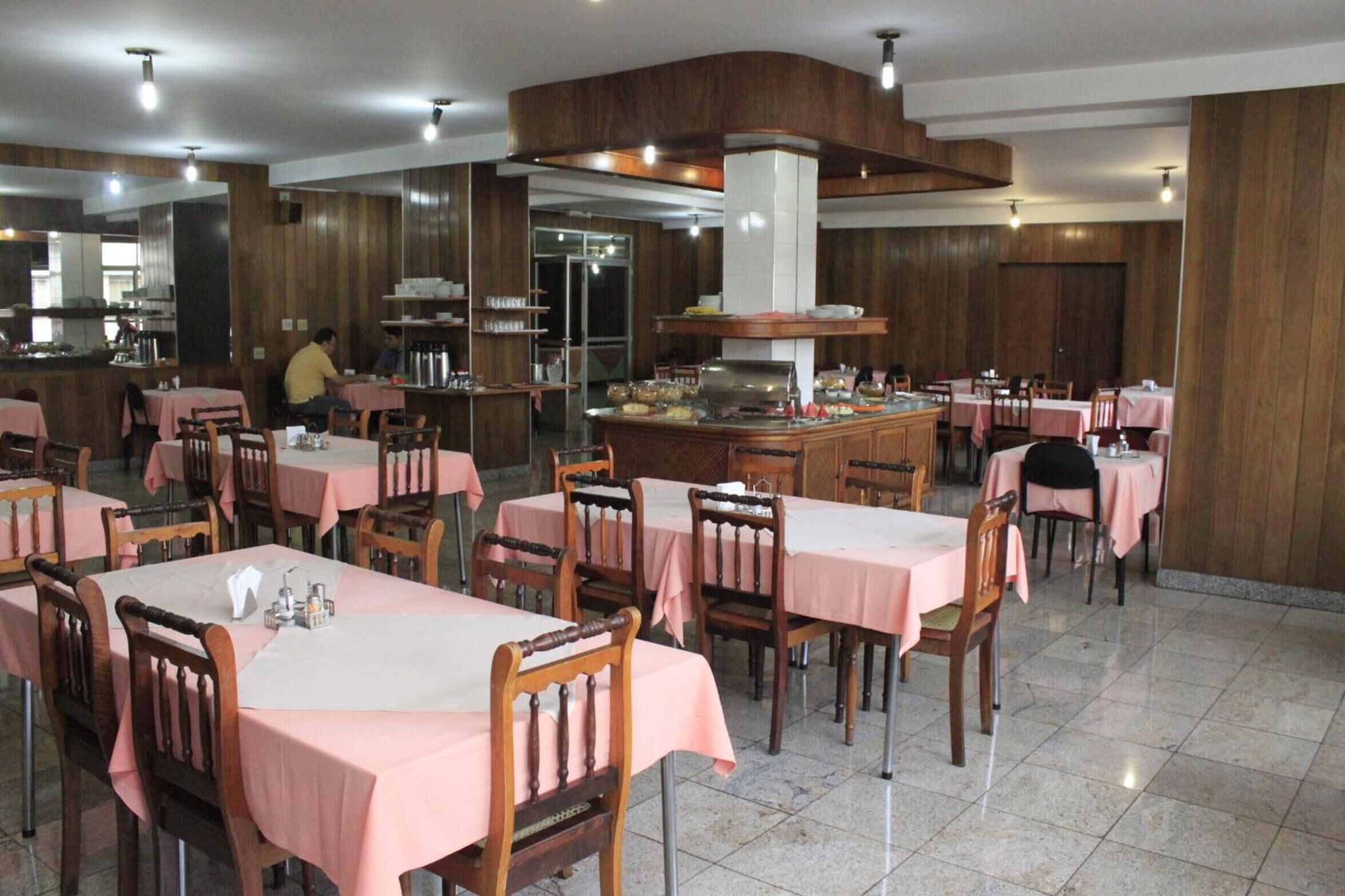 A restaurant with wooden paneling, pink tablecloths, and a central island with a stove.