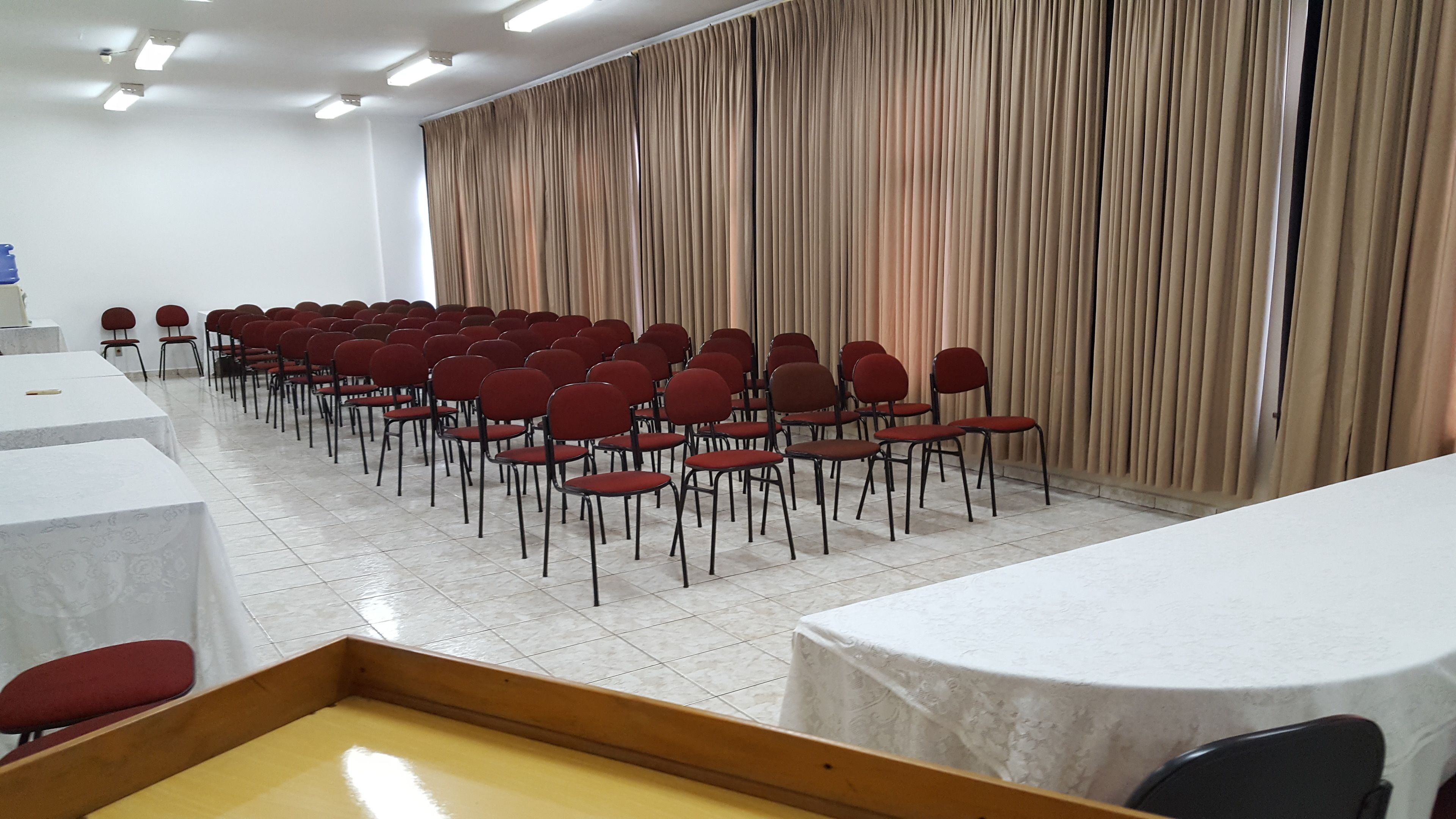Une salle de conférence avec des rangées de chaises rouges, des tables blanches et des rideaux beiges.