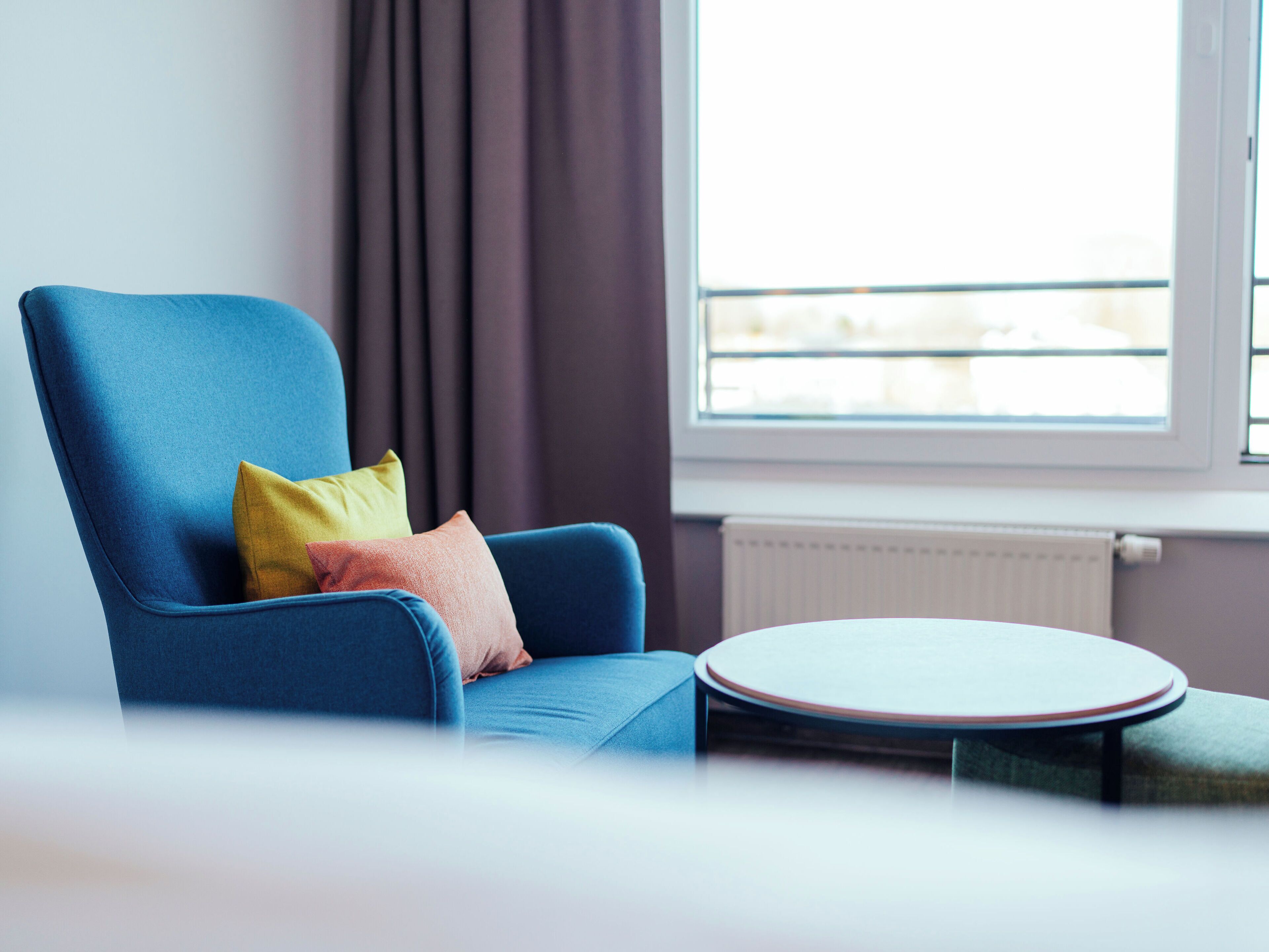 Een kamer met een blauwe fauteuil, een klein rond tafeltje, een raam met uitzicht en een radiator.