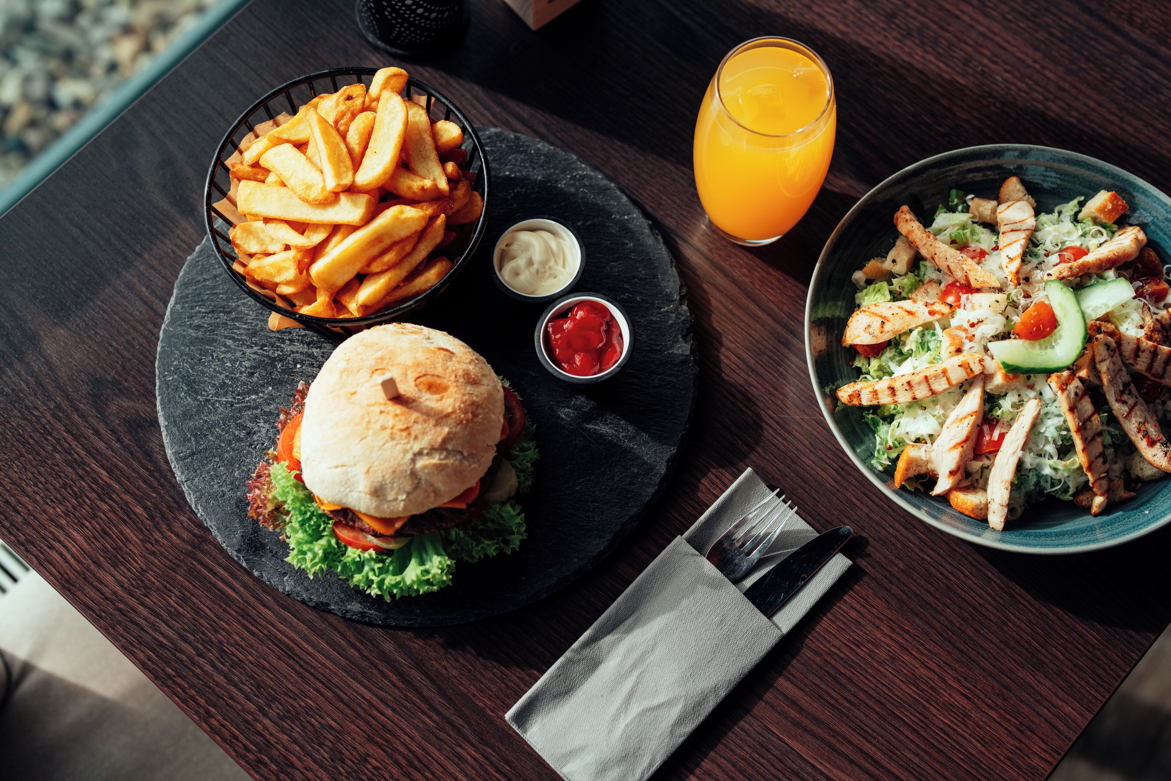 A meal with a burger, fries, and salad on a slate plate, accompanied by a glass of orange juice.