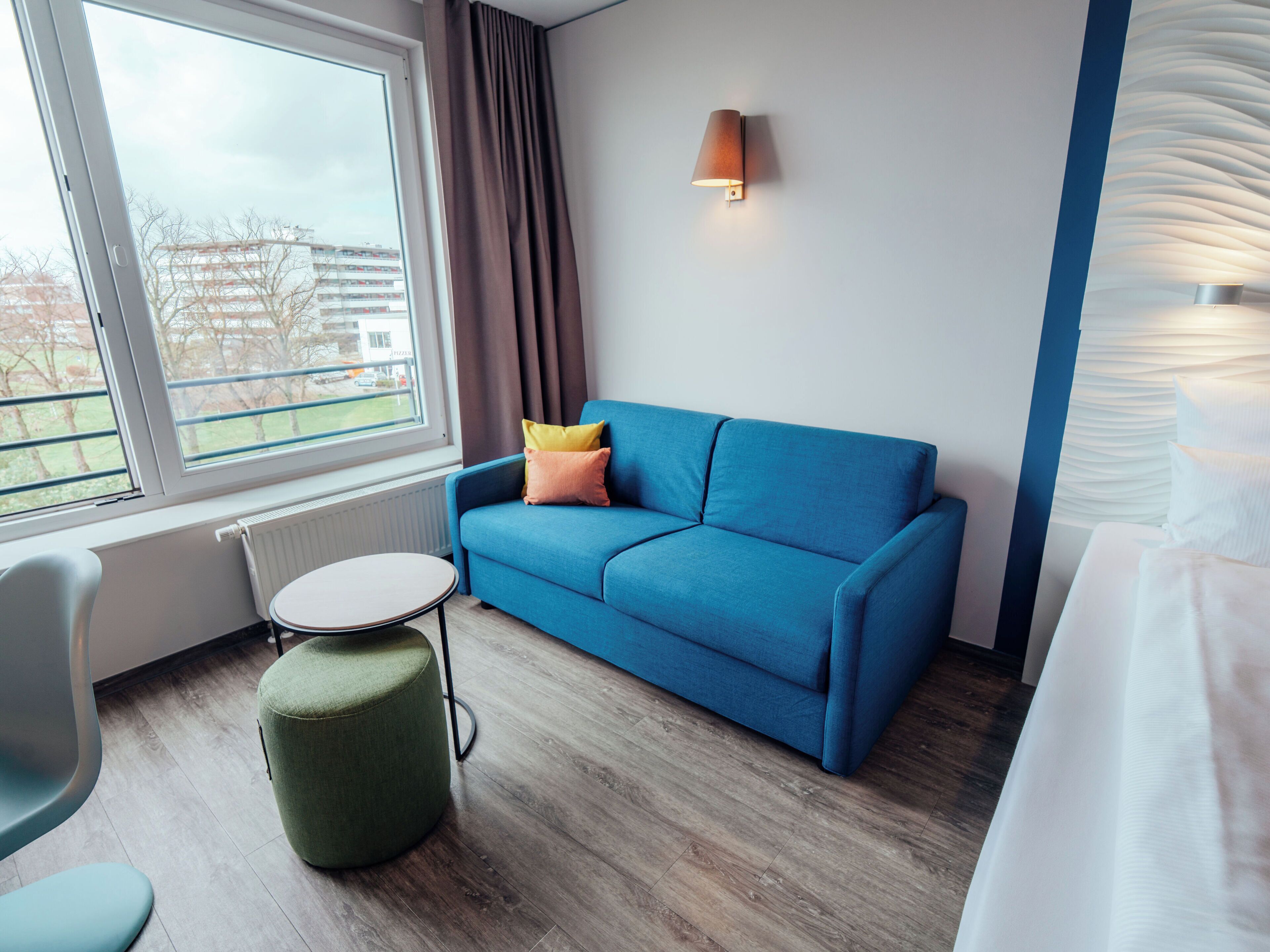 Una habitación de hotel moderna con un sofá azul, una mesita redonda pequeña, un reposapiés verde, una ventana con vistas a edificios y una lámpara fijada a la pared.
