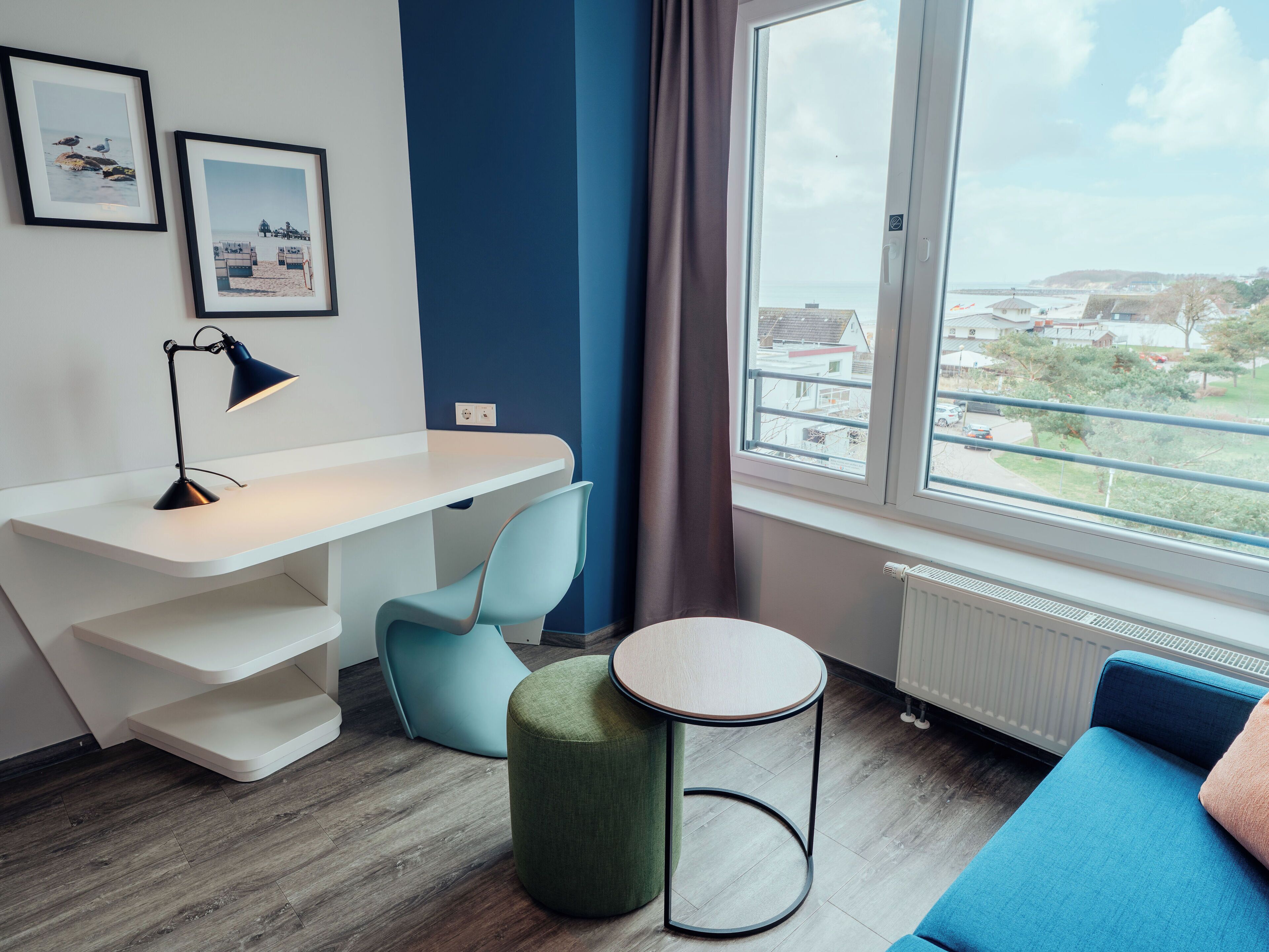 A modern room with a desk, chair, ottoman, and a view of a residential area through a large window.