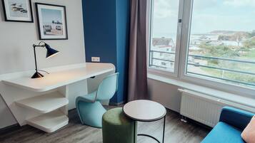 A modern room with a desk, chair, ottoman, and a view of a residential area through a large window.