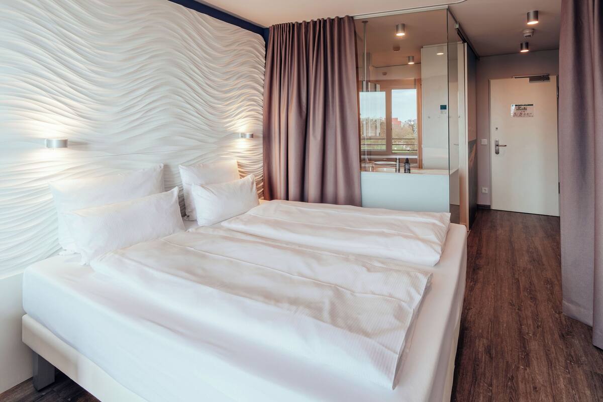 A modern hotel room with a large bed, a wall-mounted light fixture, and a view of the city through a window.
