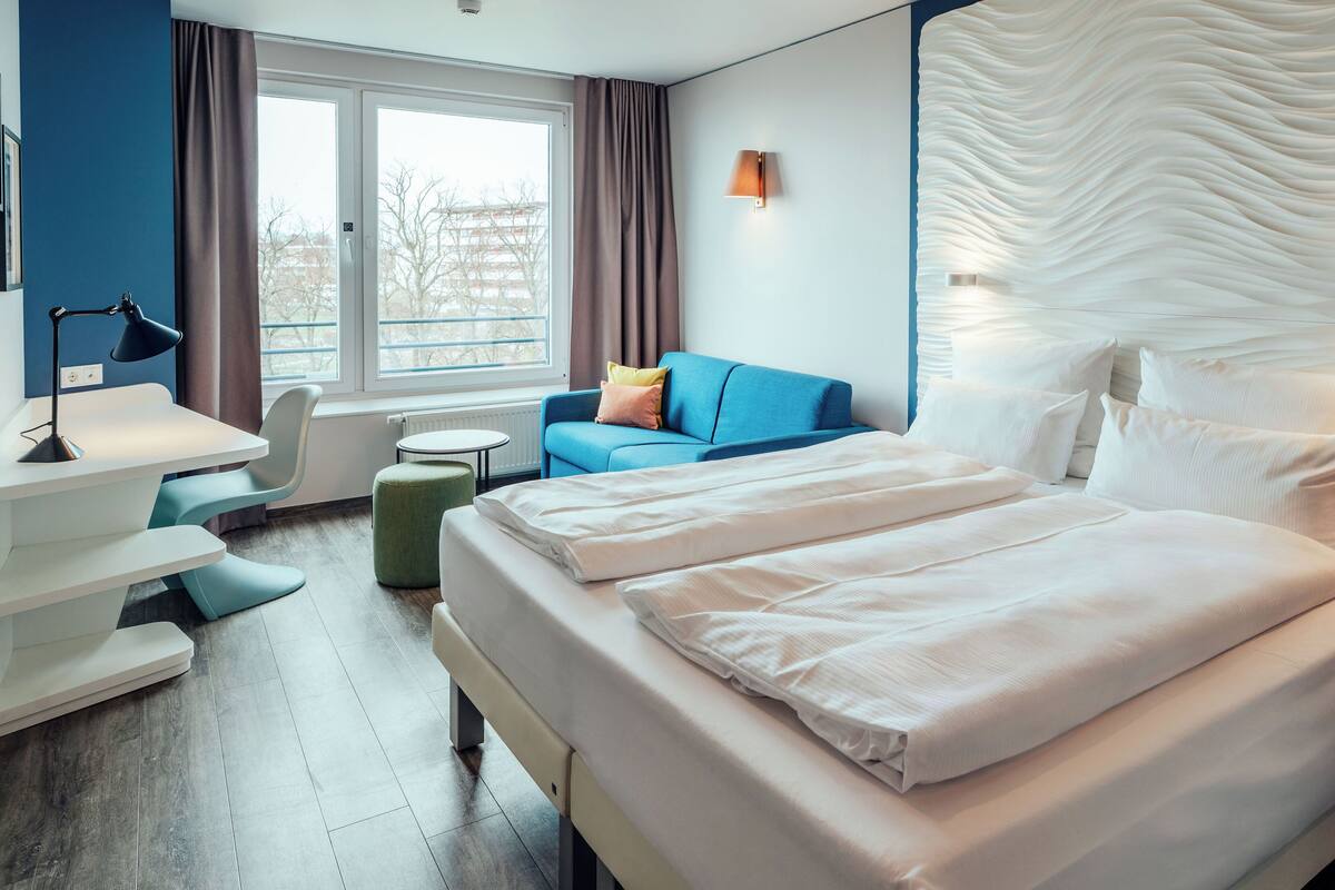 A hotel room with a large bed, a desk with a lamp, a blue sofa, and a view of the outside.