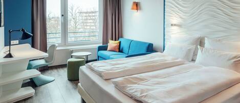 Une chambre d’hôtel avec un grand lit, un bureau avec une lampe, un canapé bleu et une vue sur l’extérieur.