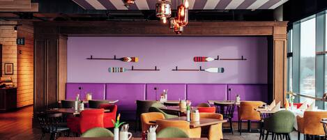A restaurant interior with wooden tables, colorful chairs, and pendant lighting.