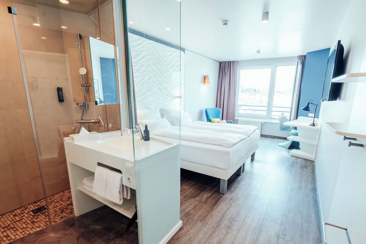 A modern hotel room with a shower, a bed, a desk, and a window.