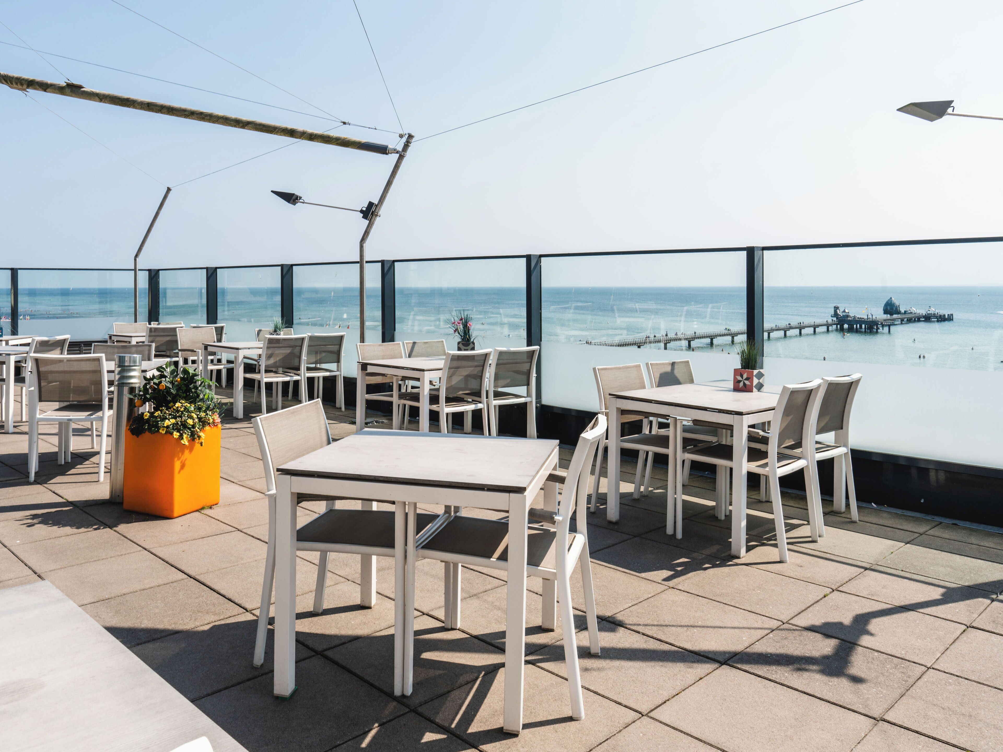 An outdoor terrace with tables and chairs, overlooking a coastal view with a pier.